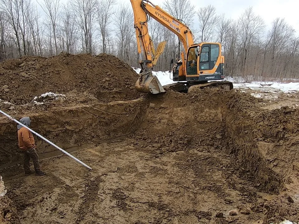 Excavator digging a basement hole on a site with patches of snow, basement excavation