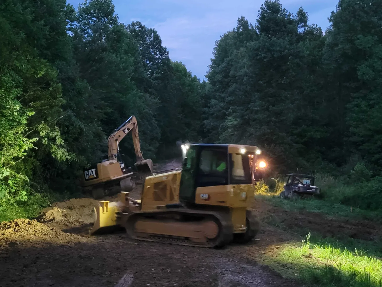 Bulldozer and excavator with work lights on a wooded site at dusk, demolition